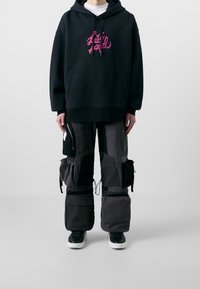 Felpa nera con cappuccio e grafica rosa, abbinata a pantaloni cargo grigi larghi con tasche e coulisse, e sneaker nere.