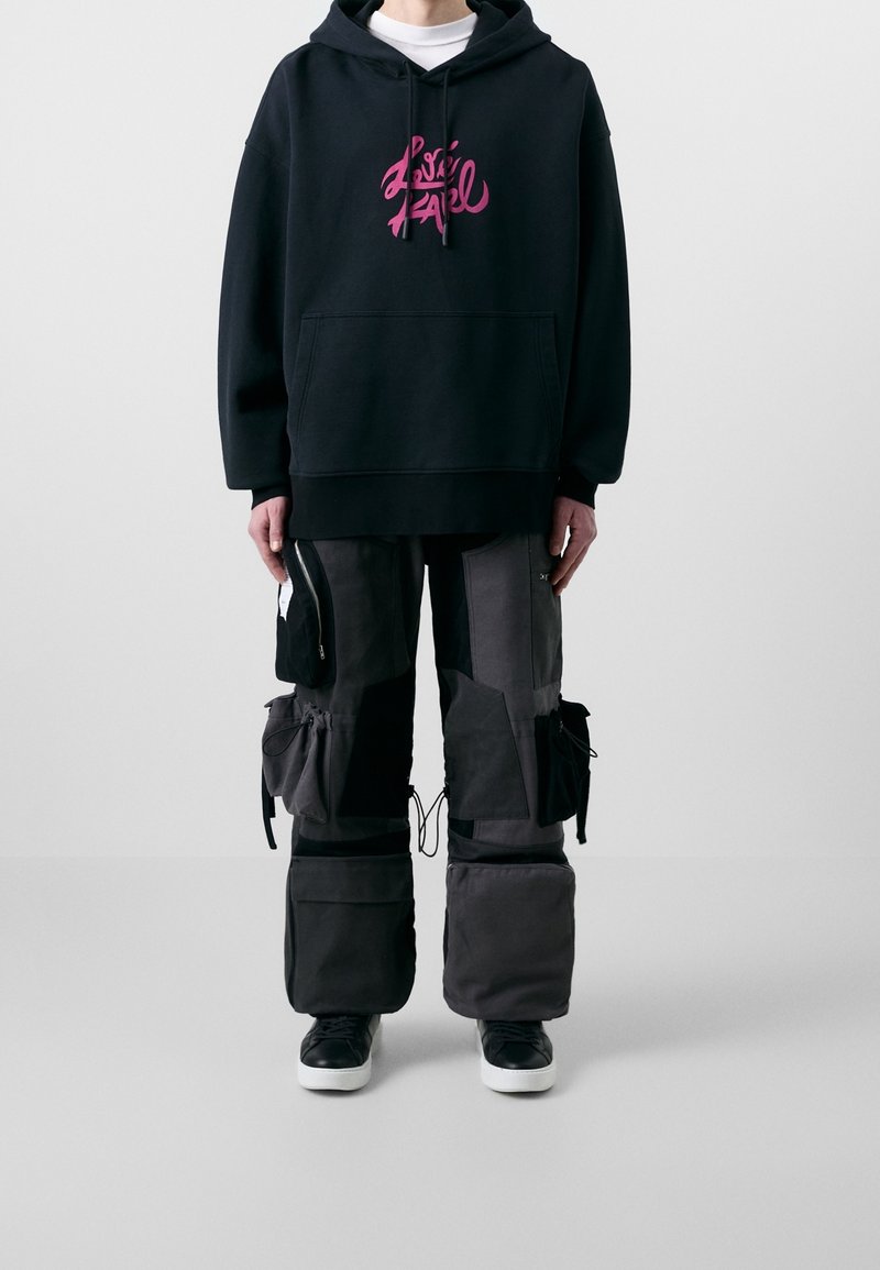 Felpa nera con cappuccio e grafica rosa, abbinata a pantaloni cargo grigi larghi con tasche e coulisse, e sneaker nere.