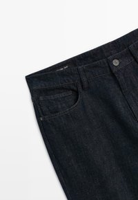 Jeans in denim scuro con vestibilità slim, dotati di cintura con bottone, cinque tasche e tessuto testurizzato. Marca visibile nella parte superiore.