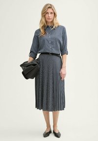 Dunkelblaues gemustertes Hemd mit hochgerollten Ärmeln, kombiniert mit einem plissierten Rock. Ein schwarzer Gürtel, eine einfarbige schwarze Tasche und flache schwarze Schuhe runden das Outfit ab.
