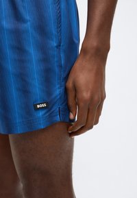 Calções de banho azuis com padrão de riscas verticais, feitos de material leve, apresentando um emblema preto com o logótipo "BOSS" e design justo ao corpo.