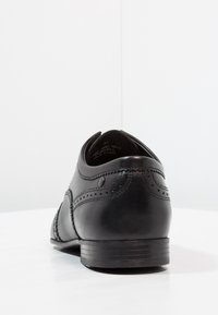 Chaussure en cuir noire avec décorations de broguing, bout arrondi, talon bas et texture lisse. Vue de dos montrant les détails de couture et le logo.