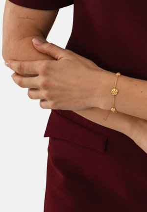 Woman wearing a gold flower charm bracelet on her left wrist, holding her right forearm, dressed in a burgundy outfit.