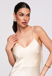 Femme aux cheveux foncés attachés en queue de cheval basse, portant une robe en satin crème avec des bretelles tressées, des boucles d'oreilles florales et des bracelets en or, regardant de côté.