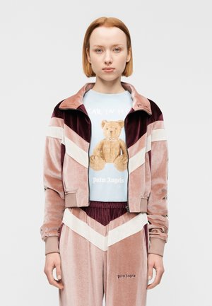 Chándal de terciopelo en tonos rosa y burdeos, con un patrón en forma de chevron blanco, combinado con una camisa azul claro que muestra un gráfico de oso.