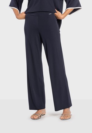 Personne portant un pantalon large ample de couleur marine, les mains sur les hanches, et des sandales plates argentées à brides, debout devant un fond uni.