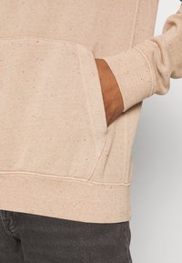 Beige sweatshirt i mjukt tyg, med en framficka, ribbade ärmar och en subtil prickig textur med fläckar av rött och svart.