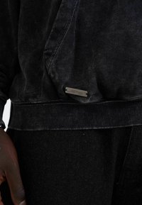 Black sweatshirt made of textured material, featuring a ribbed hem, visible stitching, and a metallic logo tag at the bottom right.