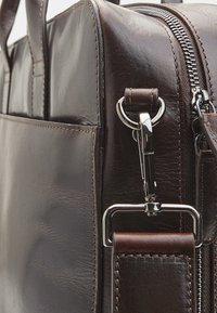 Close-up of dark brown leather bag corner showing zipper, metal clasp, and stitched strap attachment.