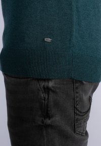 Ourlet de pull vert tricoté avec un petit logo au-dessus de la ceinture, porté avec un jean en denim noir sur le bas du torse d'une personne.