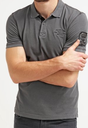 Hombre con una camisa polo gris de manga corta con texto bordado y un parche en la manga, brazos cruzados sobre el pecho.