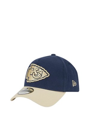 Casquette bleu marine avec une visière beige, arborant un logo KC brodé sur un patch en forme de bouclier. Fabriquée en tissu au toucher lisse.