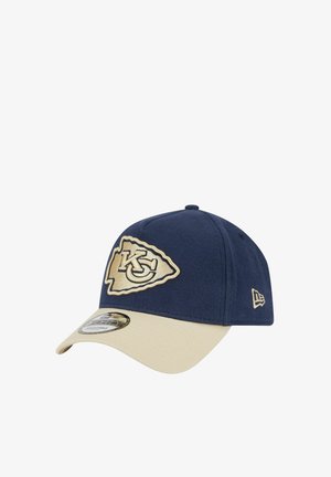 Casquette bleu marine avec une visière beige, arborant un logo KC brodé sur un patch en forme de bouclier. Fabriquée en tissu au toucher lisse.