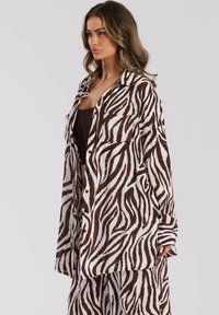 Mujer vestida con una camisa oversized de rayas de cebra marrón y blanca y pantalones a juego, con cabello suelto y ondulado, contra un fondo liso claro.