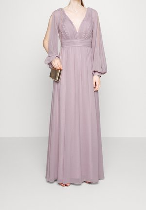Vestido largo color lavanda con escote en V profundo, cuerpo plisado y mangas abullonadas. Combinado con un pequeño bolso de mano dorado. Tejido suave y fluido.