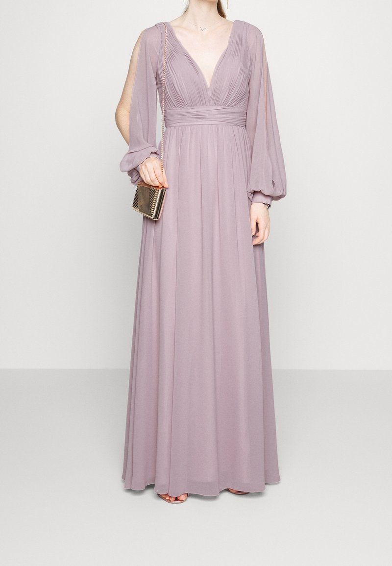 Longue robe lavande avec un décolleté en V profond, un corsage plissé et des manches bouffantes. Associée à une petite pochette dorée. Tissu lisse et fluide.