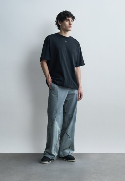 Νεαρός άνδρας με σγουρά μαλλιά, φορώντας μια φαρδιά μαύρη μπλούζα και φαρδιά ανοιχτόγρια τζιν, στέκεται μπροστά σε έναν μονόχρωμο ανοιχτό τοίχο.