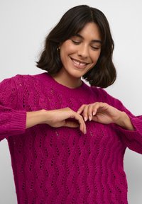 Maglione lavorato a maglia fucsia con motivo traforato, girocollo e polsini a coste. Il modello mostra una vestibilità rilassata con movimenti delle mani visibili.