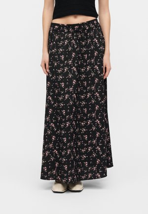 Kvinde iført en sort blomster maxi nederdel med pink blomster, elastisk talje, sort crop top og beige sko med firkantet tå.
