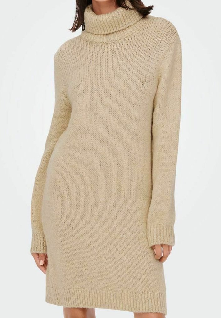 Robe-pull beige à col roulé en maille à manches longues portée par une personne aux cheveux bruns, debout devant un fond blanc uni.