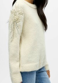 Heller beiger Pullover aus weichem, strukturiertem Stoff. Er verfügt über ein dekoratives Design mit Perlen und Pailletten auf einem Ärmel.