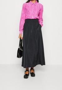 Camicia a righe rosa a maniche lunghe con bottoni, abbinata a una gonna maxi in satin nera. Tiene in mano una borsa lucida nera; indossa scarpe platform nere.