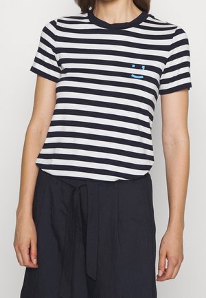 Frau trägt ein schwarz-weiß gestreiftes T-Shirt mit horizontalen Streifen und einem kleinen blauen Smiley auf der Brust, kombiniert mit schwarzen Hosen mit Bindegürtel.