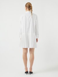 Robe chemise plissée blanche à manches longues, avec un col et un ourlet évasé. Associée à des chaussures à talons hauts noires. Texture de tissu lisse.