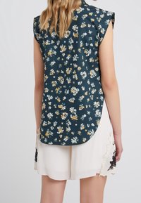 Blusa senza maniche floreale in verde scuro, con orlo curvo e vari piccoli fiori bianchi e gialli, abbinata a pantaloni corti color crema chiaro.
