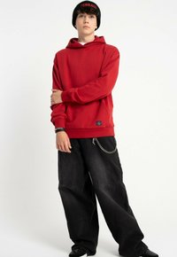 Czerwony hoodie z kieszenią kangurzą i metką, w połączeniu z czarnymi spodniami o szerokim kroju z przeszyciami z przodu i akcentem w postaci łańcuszka.