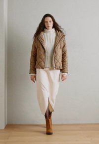 Doudoune matelassée brun, pull en maille blanc, jupe blanche avec fente, et bottes brunes à lacets. Texture lisse, design superposé, couleurs neutres.