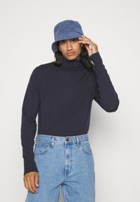 Maglione a collo alto blu navy con maniche lunghe, abbinato a jeans di denim chiaro a vita alta. Un cappello a secchiello in velluto a coste blu completa il look.