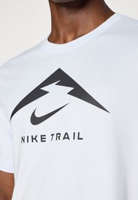 Bílé tričko s krátkým rukávem, na přední straně velké černé logo Nike a nápis "NIKE TRAIL", vyrobené z měkkého bavlněného materiálu.