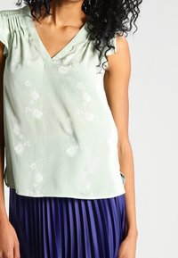 Blusa senza maniche di colore verde chiaro con scollo a V e motivo floreale. Realizzata in un tessuto testurizzato, presenta delle pieghe sulle spalle.