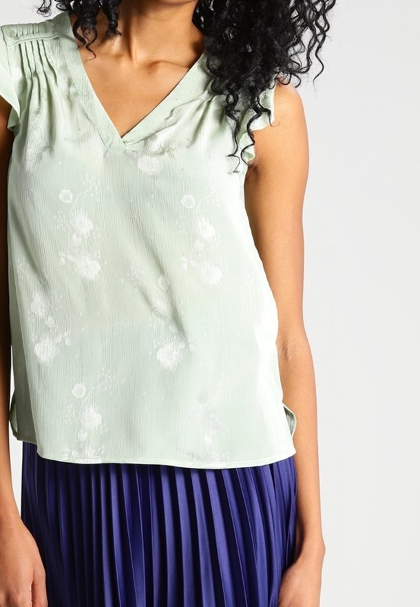 Blusa senza maniche di colore verde chiaro con scollo a V e motivo floreale. Realizzata in un tessuto testurizzato, presenta delle pieghe sulle spalle.