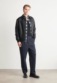 Zwarte leren jas, geruite shirt met blauwe en groene tinten, zwart tanktop, marineblauwe broek, zwarte schoenen. Soepele textuur, casual ontwerp.