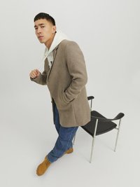 Brauner Wollmantel über grauem Hoodie, blaue Jeans und braunen Stiefeln. Schwarzer Stuhl mit verchromten Beinen im Hintergrund. Einfaches, modernes Design.