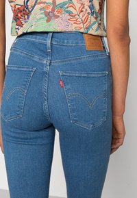 Persona vistiendo jeans Levi's azules y una blusa floral colorida, mostrada desde atrás con los bolsillos traseros visibles y las etiquetas de la marca.