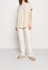 Beige proužkované triko s krátkým rukávem, spárované s krémovými kalhotami rovného střihu a světle hnědými sandály; minimalistický design a měkká textura.