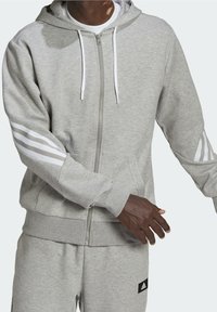 Felpa con cappuccio grigia con zip, realizzata in tessuto morbido, con cordoncini bianchi e tre strisce bianche sulle maniche. Include una tasca frontale.