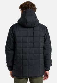 Hombre con una chaqueta acolchada con capucha negra y pantalones verdes, visto desde atrás sobre un fondo claro.