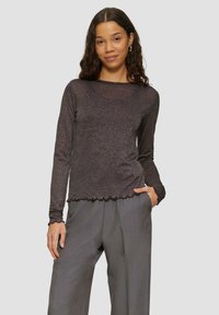 Langärmliges Oberteil aus dunklem Metallgewebe mit gewelltem Saum und figurbetontem Design. Kombiniert mit lockeren grauen Hosen.