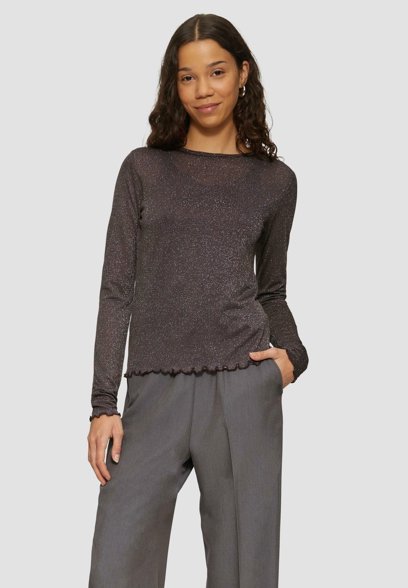 Langärmliges Oberteil aus dunklem Metallgewebe mit gewelltem Saum und figurbetontem Design. Kombiniert mit lockeren grauen Hosen.