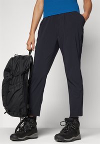 Osoba v černých horských botách a černých kalhotách, držící černý batoh za horní rukojeť.