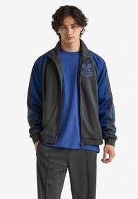 Giacca sportiva nera e blu con zip intera, dotata di un logo da tennis e maniche a contrasto, abbinata a una maglietta blu e pantaloni scuri.