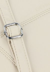 Detalle de bolso de piel crema con un hebilla cuadrada plateada. Presenta costuras visibles y una textura suave.