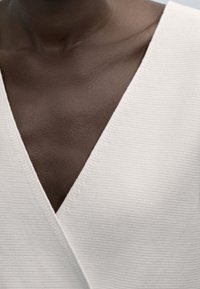 Zbliżenie na ciemną skórę szyi i górnej części klatki piersiowej, ubraną w białą, teksturowaną bluzkę z dekoltem w serek o żebrowanym wzorze.