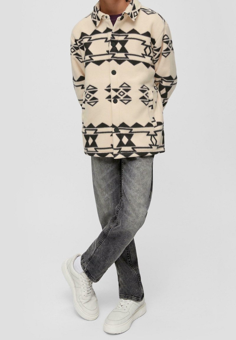 Personne portant une veste crème avec des motifs géométriques noirs, un jean gris foncé, des baskets blanches, debout jambes croisées sur un fond clair.