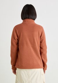 Rdzawoczerwony polarowy kurtka z wysokim kołnierzem, fakturowana tkanina i obniżone szwy ramion, noszona z jasną spódnicą.