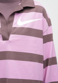 Πολυτελές ριγέ ρουμπι στυλ μπλουζάκι σε μωβ και καφέ χρώμα, με το λογότυπο Nike σε λευκό χρώμα στο στήθος και με μικρό γιακά σε σχήμα V.